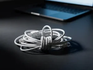 Tangled white ethernet cables coiled in a knot on a dark desk, with a half-open laptop casting blue light in the background.